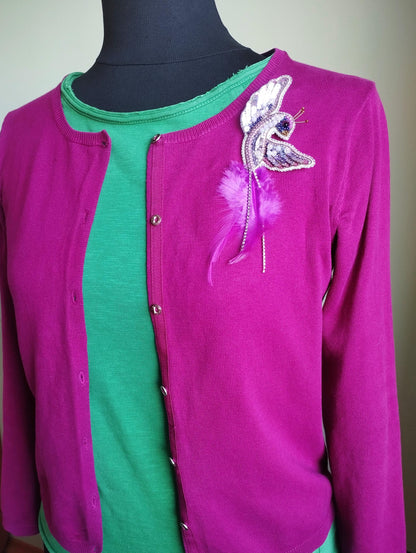 A mannequin wearing a pink cardigan with a green shirt underneath, and a purple feather pin on the front.