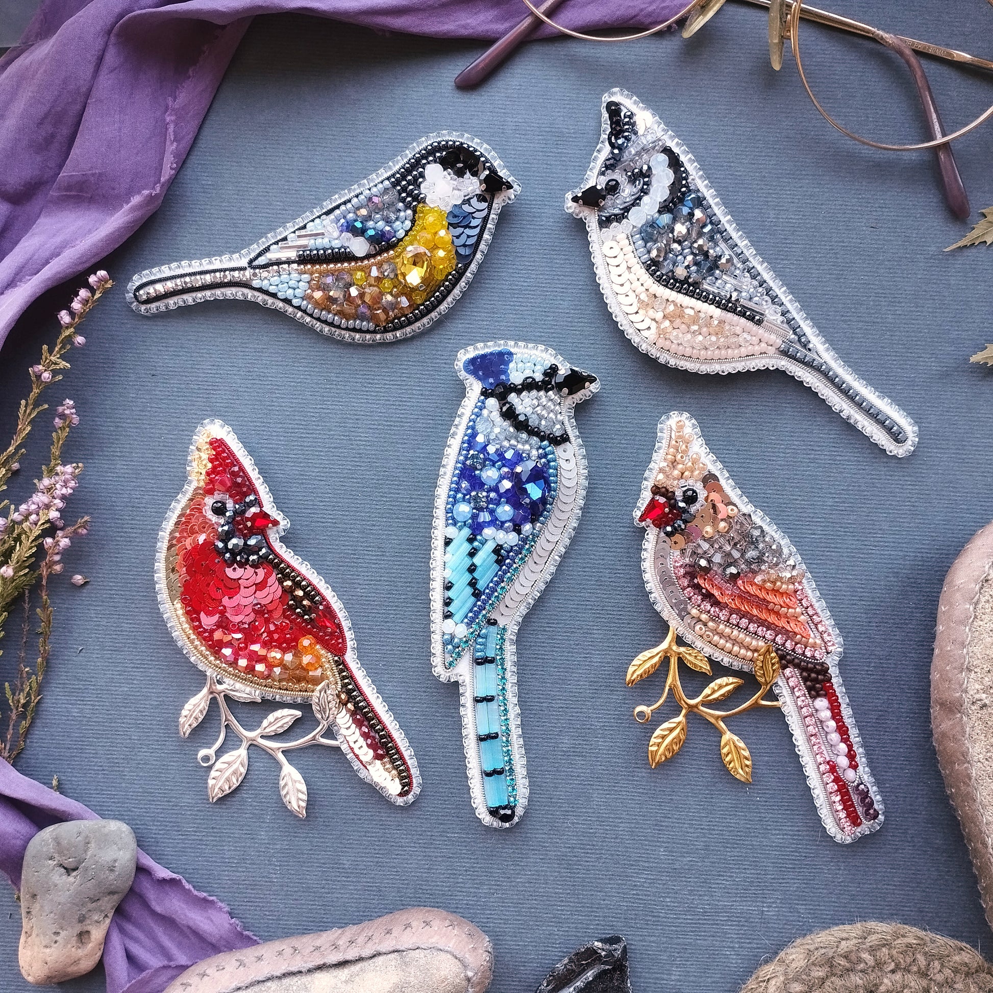 five colorful bird brooches, each with unique designs and patterns, arranged on a blue background.