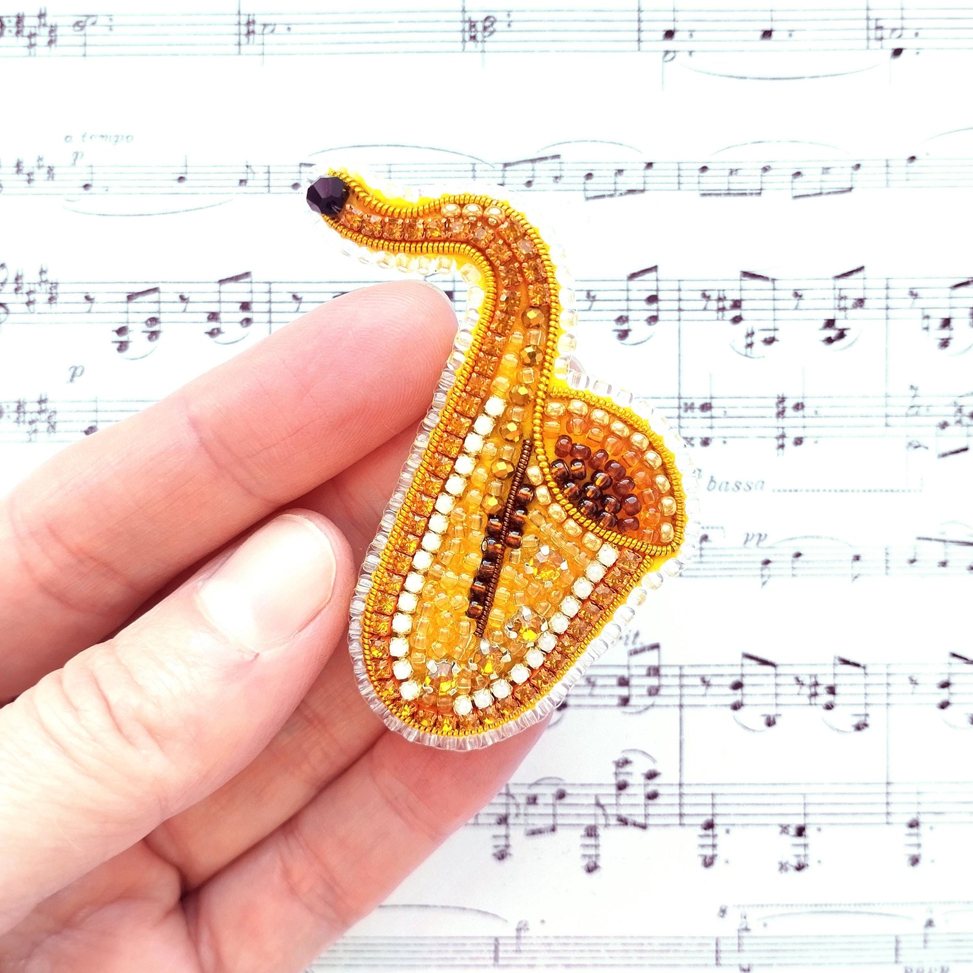 A hand is holding a small, intricately designed beaded object resembling a saxophone, with sheet music visible in the background.