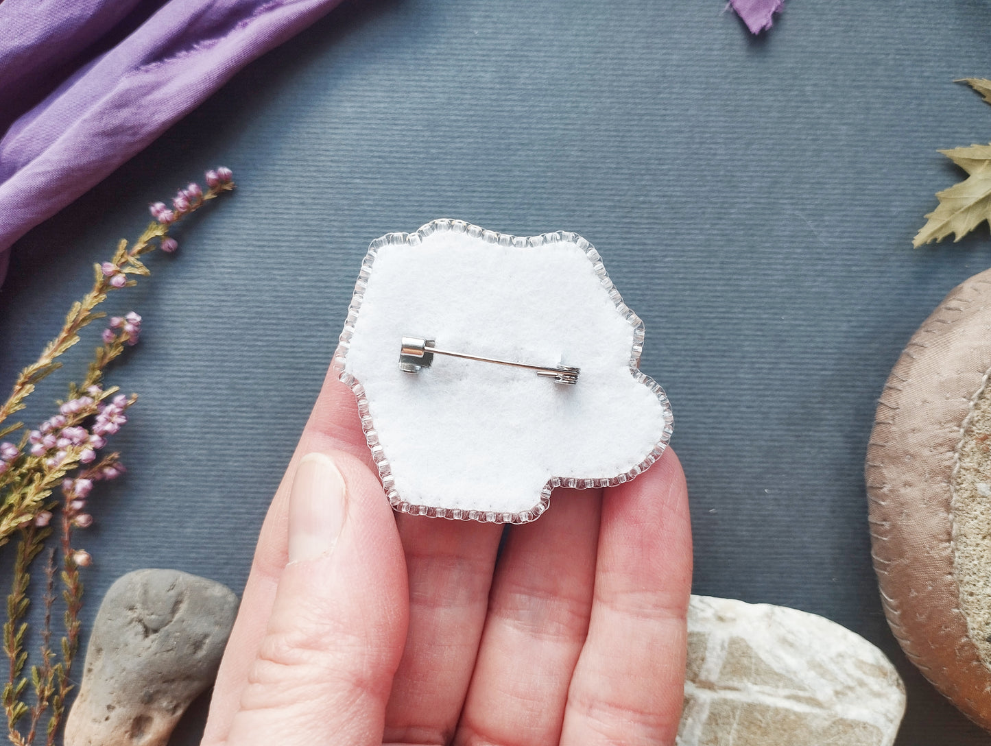A hand holding a white pin cushion with a silver pin attached to it, surrounded by various natural elements such as flowers, rocks, and leaves.