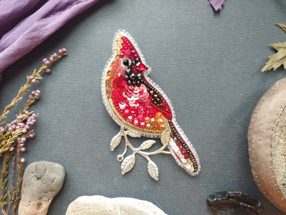 a vibrant, hand-beaded bird brooch with a red and gold color scheme, adorned with sequins and leaves, placed on a gray background.