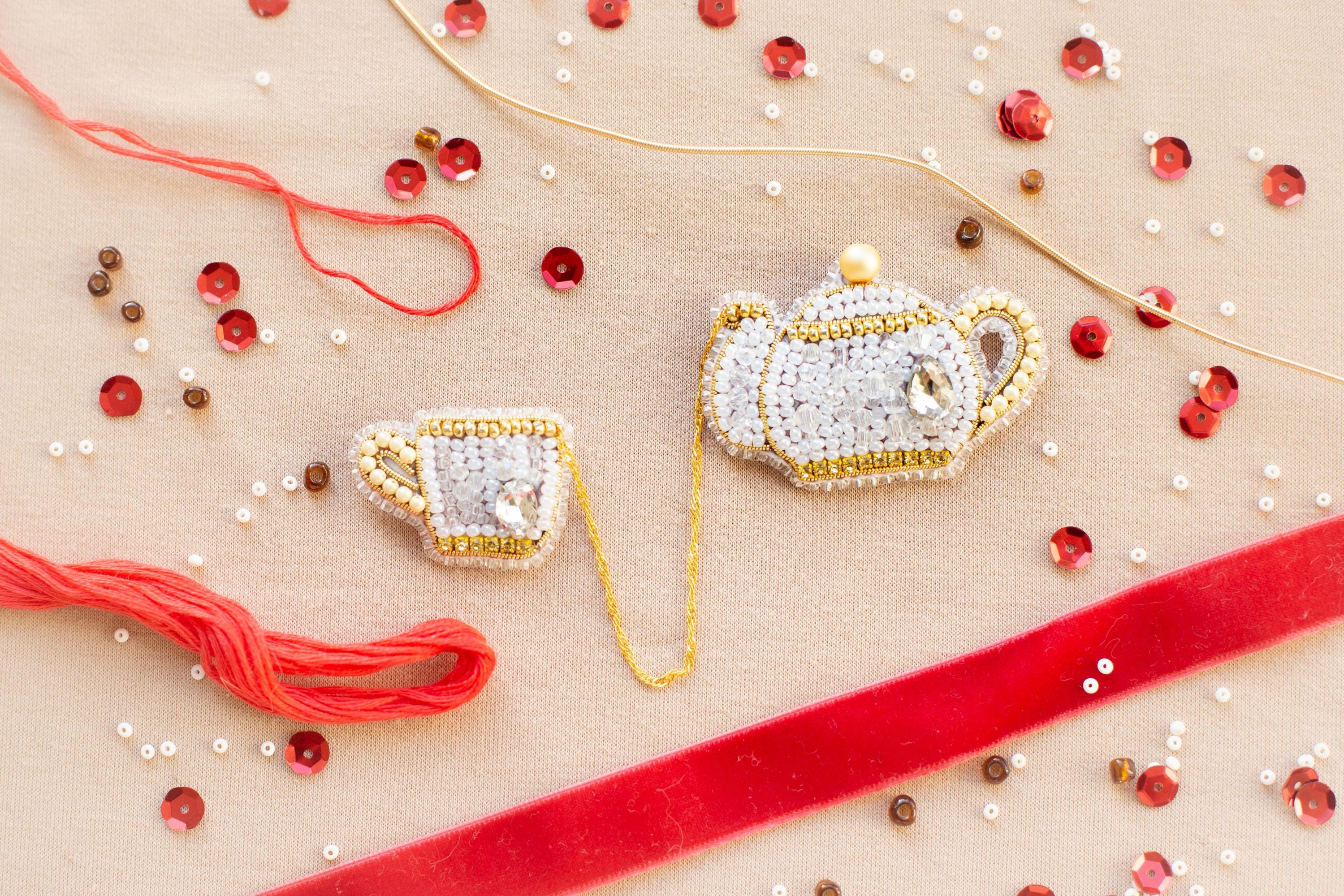 a close-up view of a beaded tea set, including a teapot, a cup, and a saucer, all intricately crafted with beads and strings. The tea set is surrounded by various beads, including red, gold, and white ones, as well as a red ribbon, creating a visually appealing and detailed arrangement.