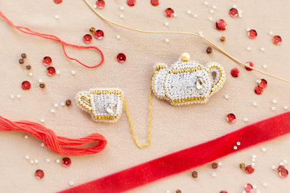 a close-up view of a beaded tea set, including a teapot, a cup, and a saucer, all intricately crafted with beads and strings. The tea set is surrounded by various beads, including red, gold, and white ones, as well as a red ribbon, creating a visually appealing and detailed arrangement.