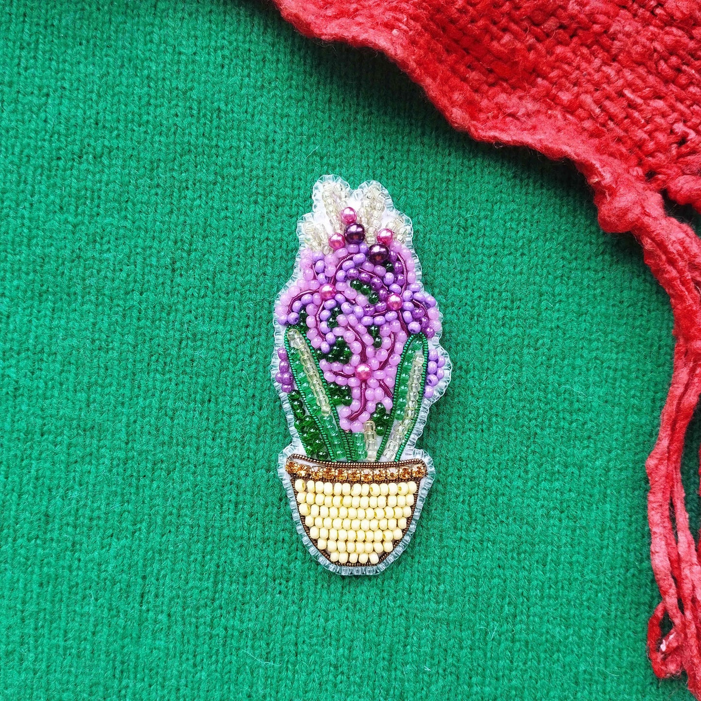 A beaded brooch in the shape of a flower is displayed on a green fabric, with a red fabric piece on the right side.