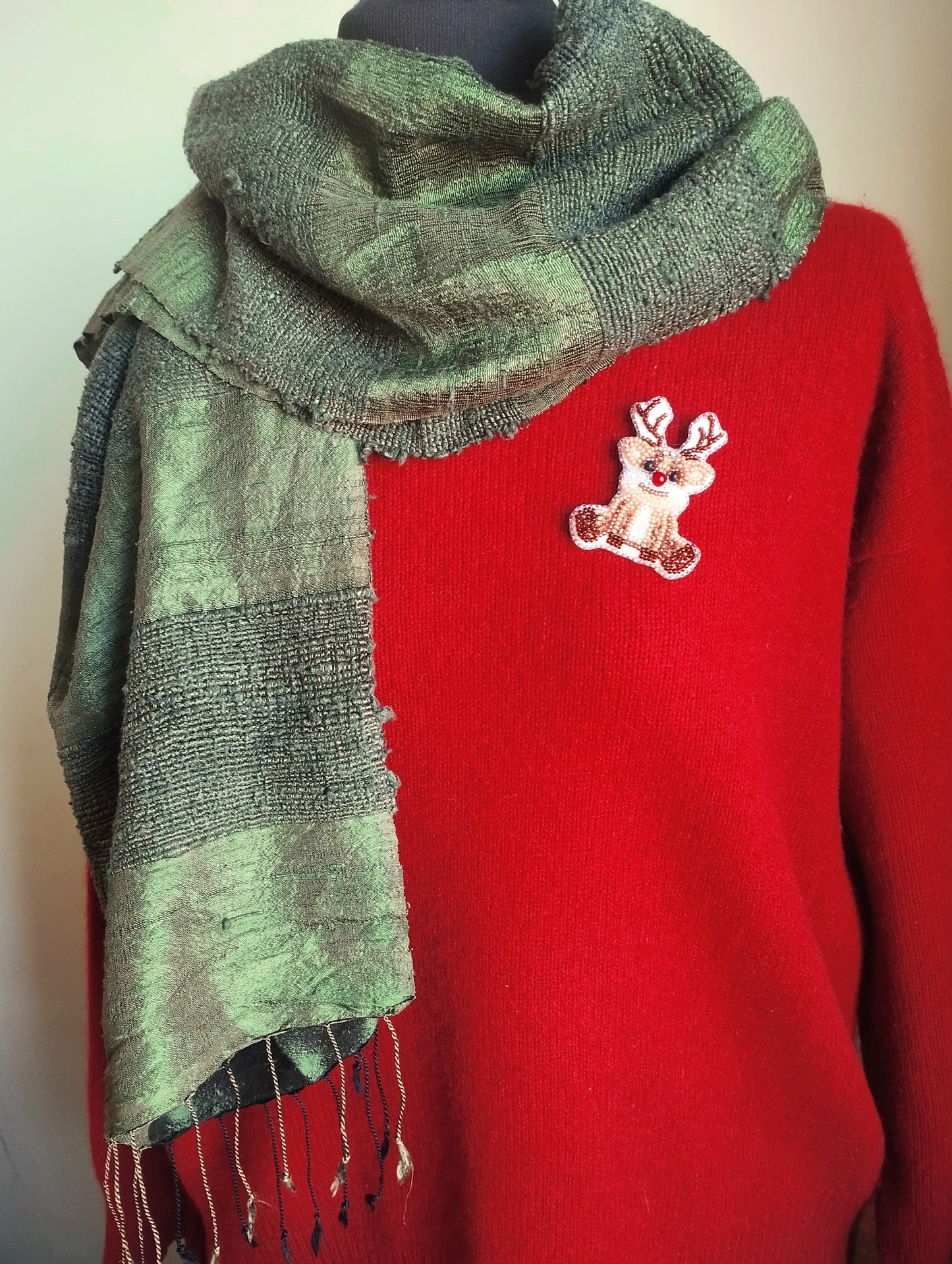A mannequin is wearing a red sweater with a green and gray scarf draped over it, and a small white patch with a red deer embroidered on the sweater.
