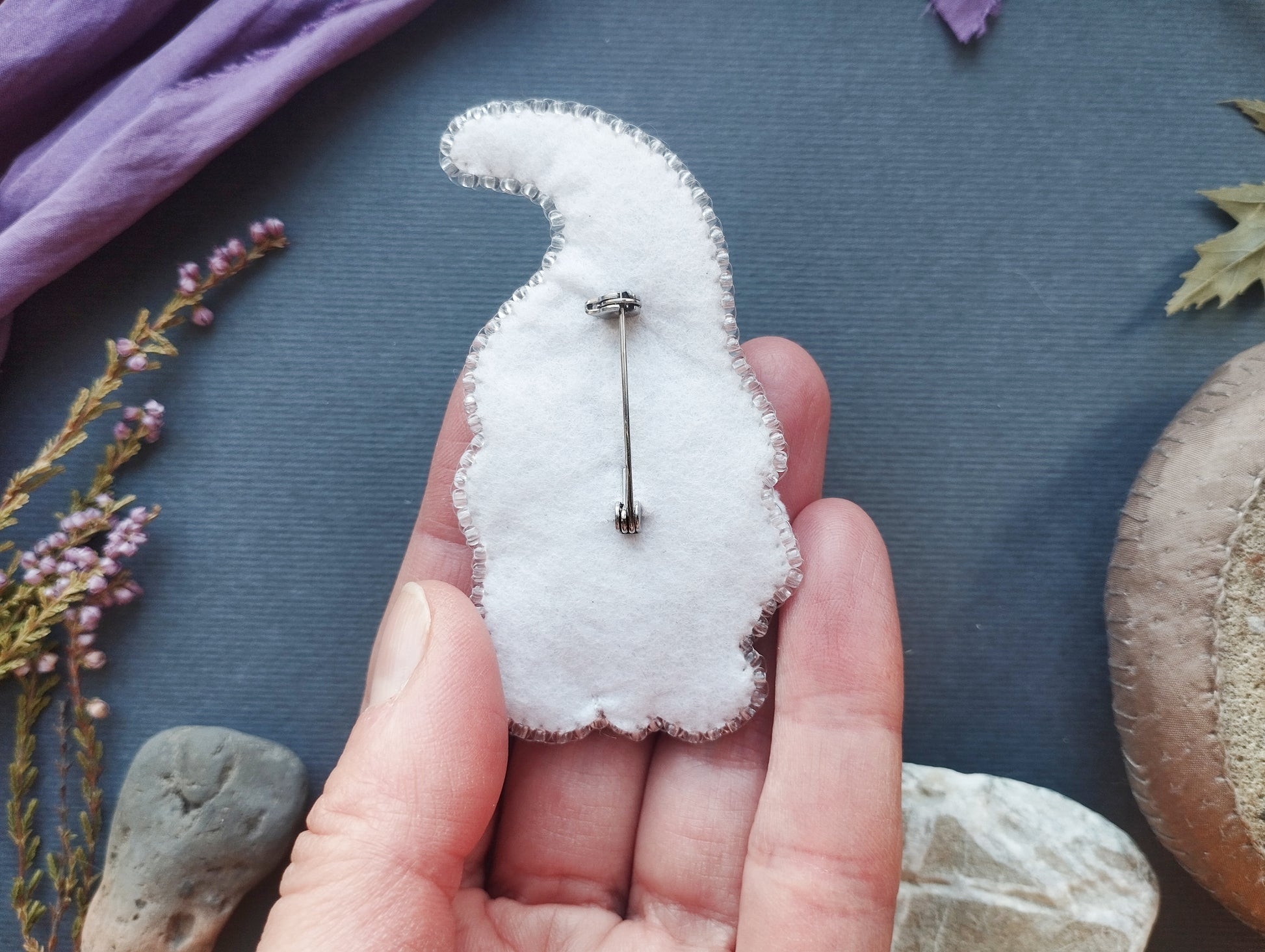 A hand holding a white, hand-sewn pin or badge against a blue background with various natural elements, such as flowers and rocks.
