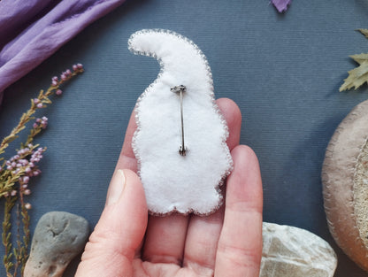A hand holding a white, hand-sewn pin or badge against a blue background with various natural elements, such as flowers and rocks.