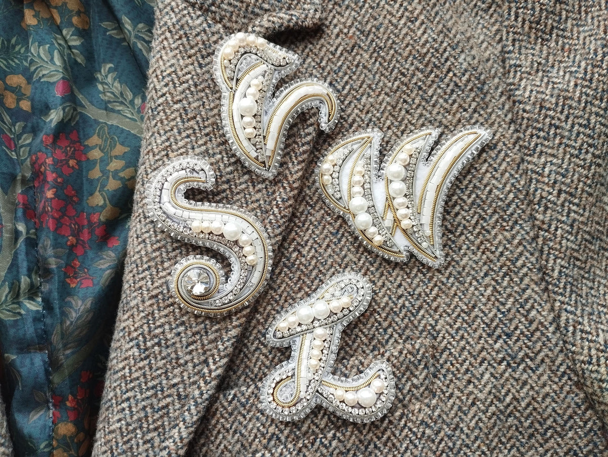 a close-up view of a grey tweed jacket with several decorative embellishments, including a large letter "S" and a smaller letter "W" made of pearls and rhinestones.