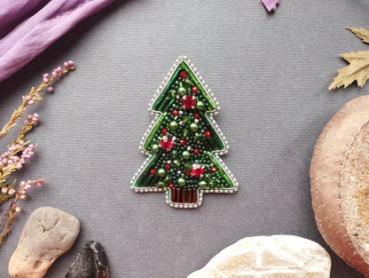 A decorative Christmas tree made of beads and other embellishments is displayed on a gray background, surrounded by various natural elements such as leaves, flowers, and rocks.