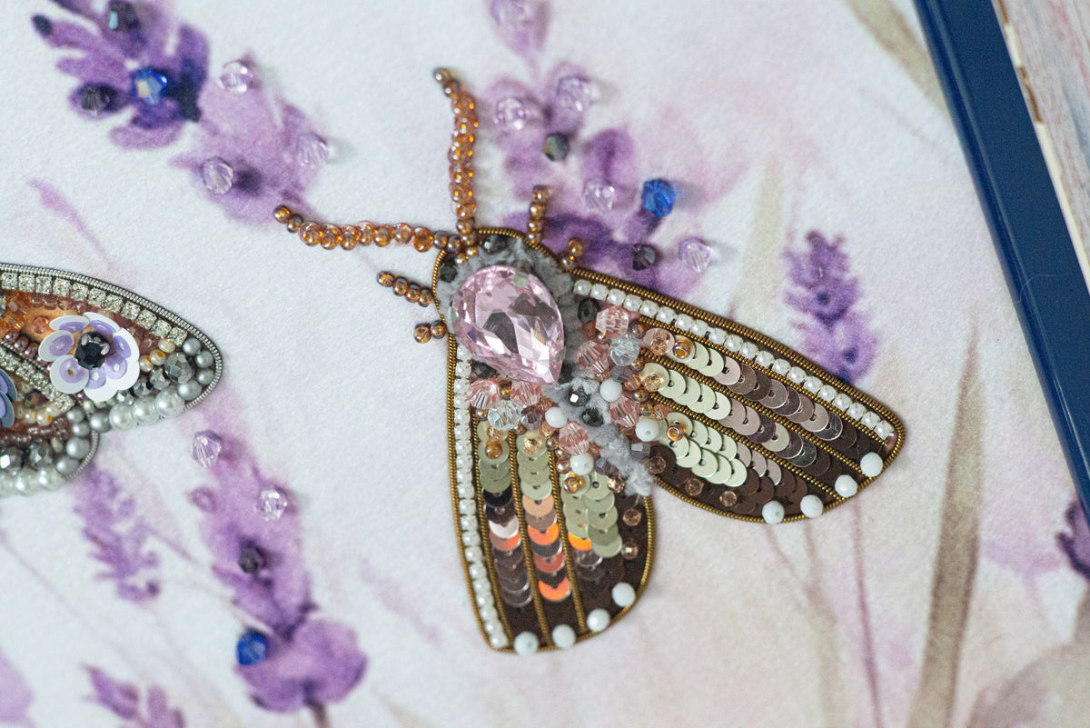 a close-up view of a butterfly-shaped embroidered piece adorned with various colorful beads and sequins, set against a background of purple flowers.