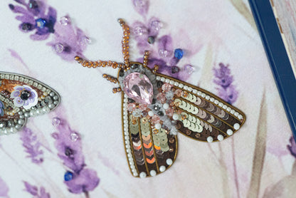 a close-up view of a butterfly-shaped embroidered piece adorned with various colorful beads and sequins, set against a background of purple flowers.