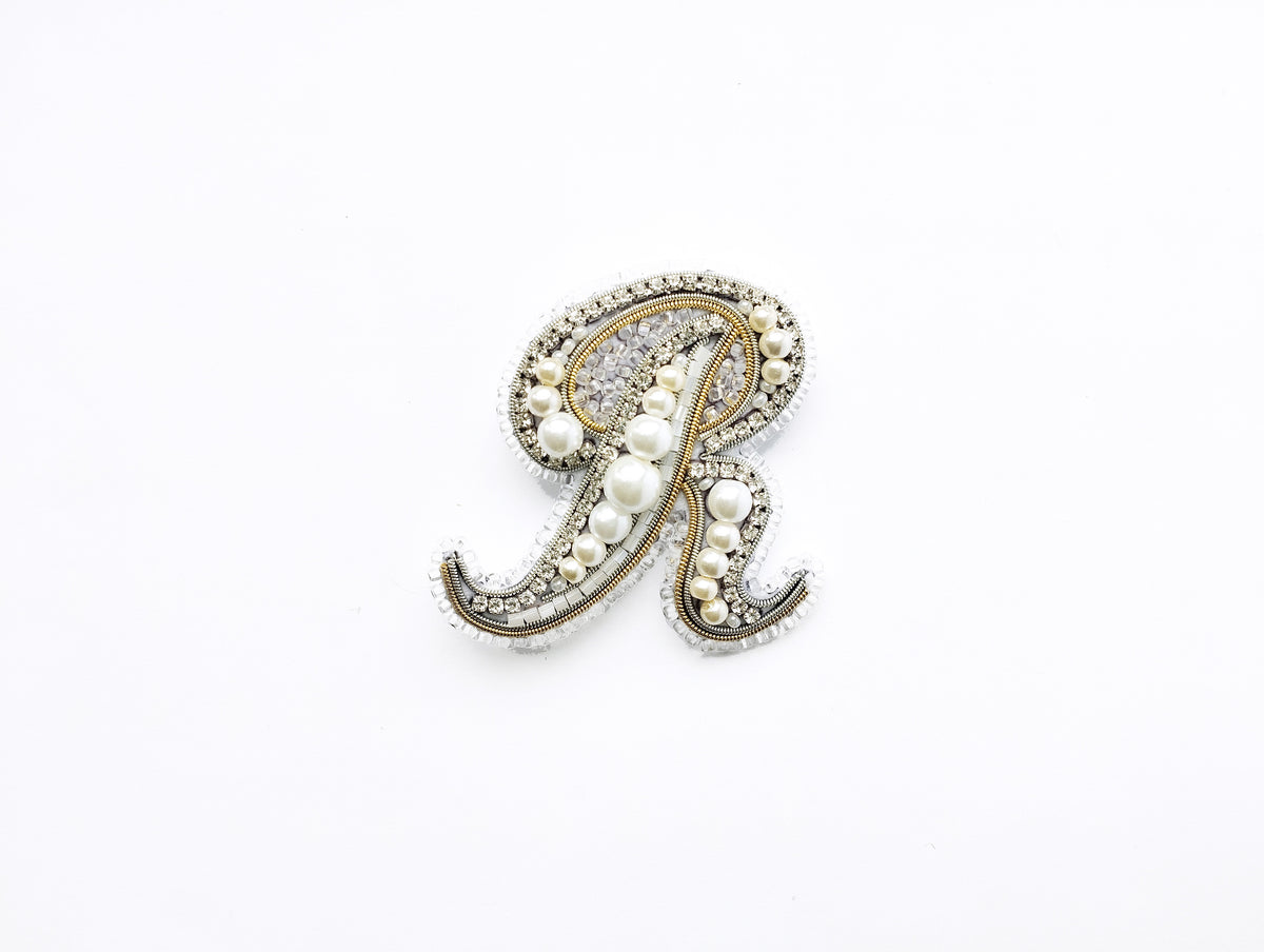 A decorative letter 'R' made of pearls and jewels against a white background.
