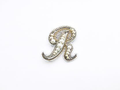 A decorative letter 'R' made of pearls and jewels against a white background.