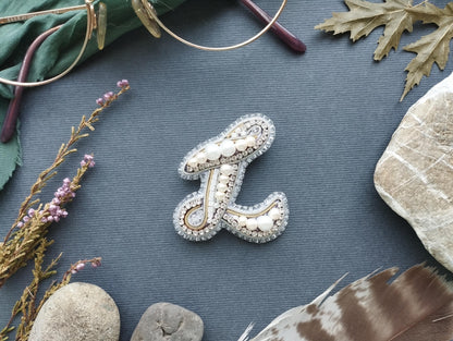 a close-up of a decorative letter 'L' made of beads, pearls, and other embellishments, surrounded by various natural elements such as feathers, leaves, and rocks.