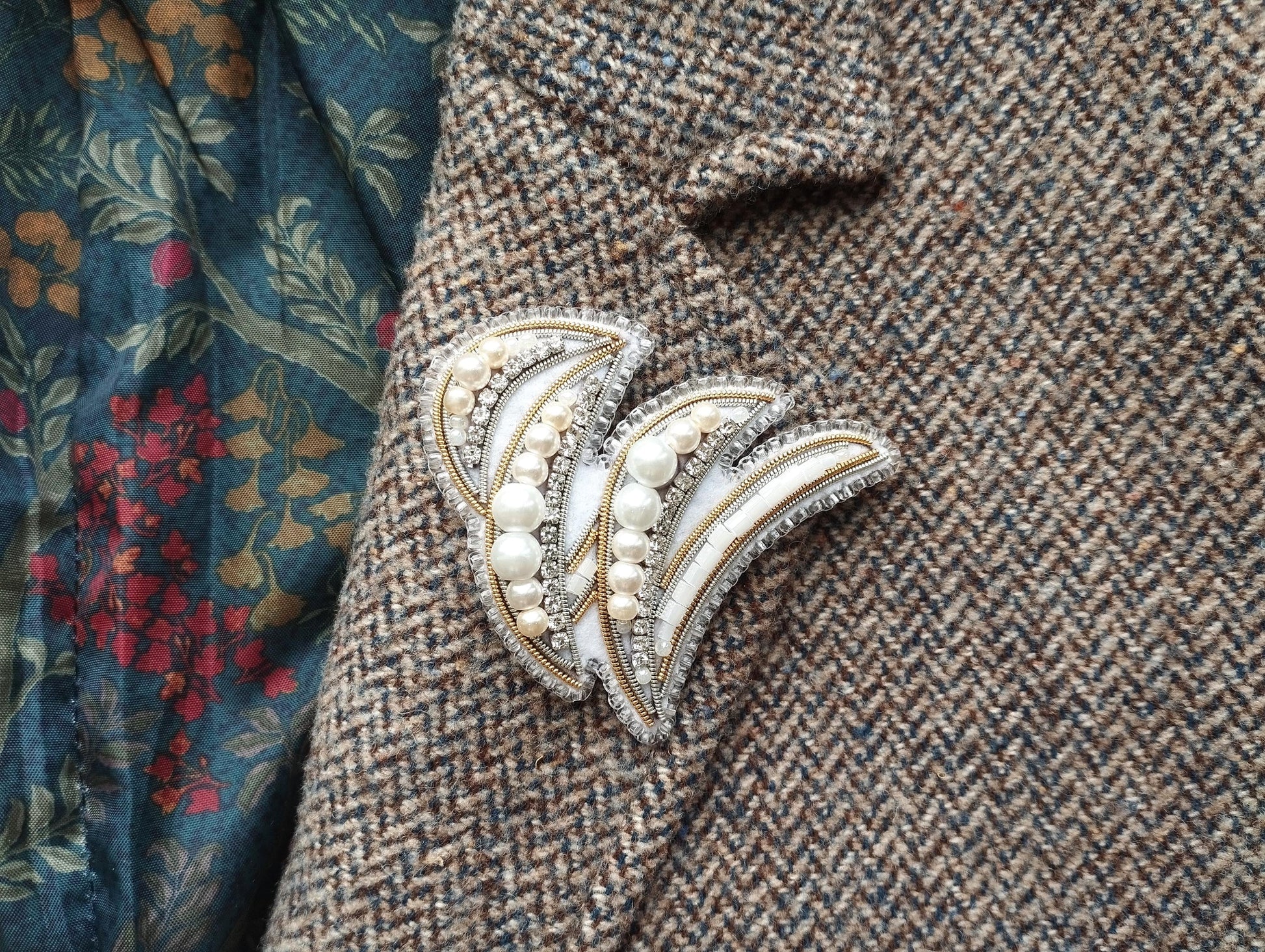 a close-up view of a decorative brooch or pin with pearls and a floral design, placed on a tweed jacket or fabric with a floral pattern.