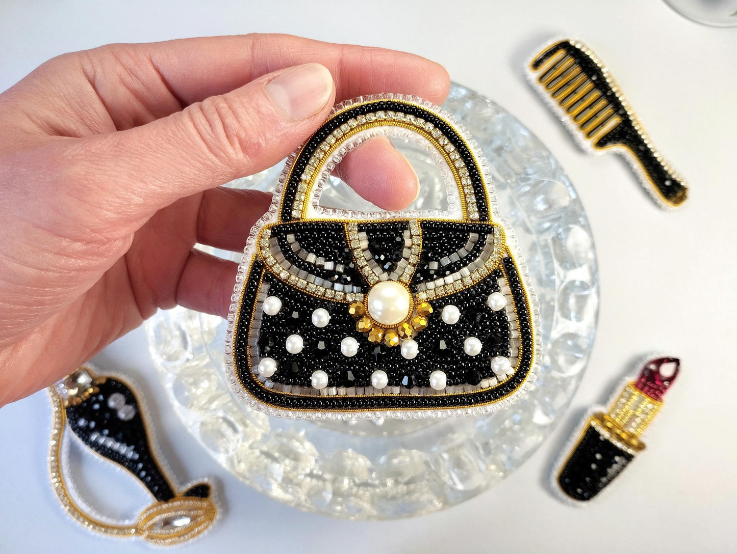 A hand holding a handbag-shaped embroidered object with a pearl at the center, surrounded by other decorative items such as a comb and a lipstick.