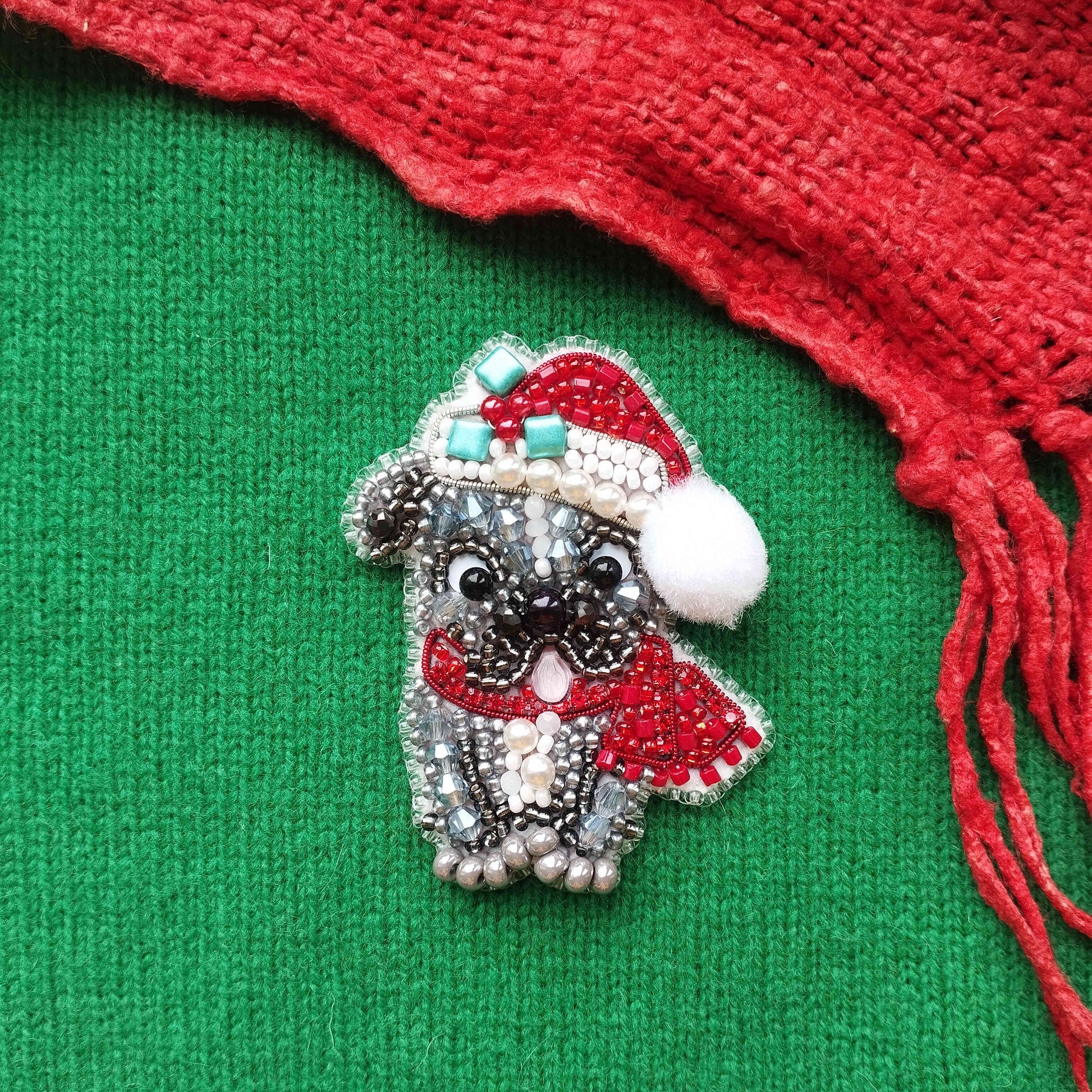 A decorative beaded brooch in the shape of a dog wearing a Santa hat and scarf, placed on a green fabric background with a red fringe.