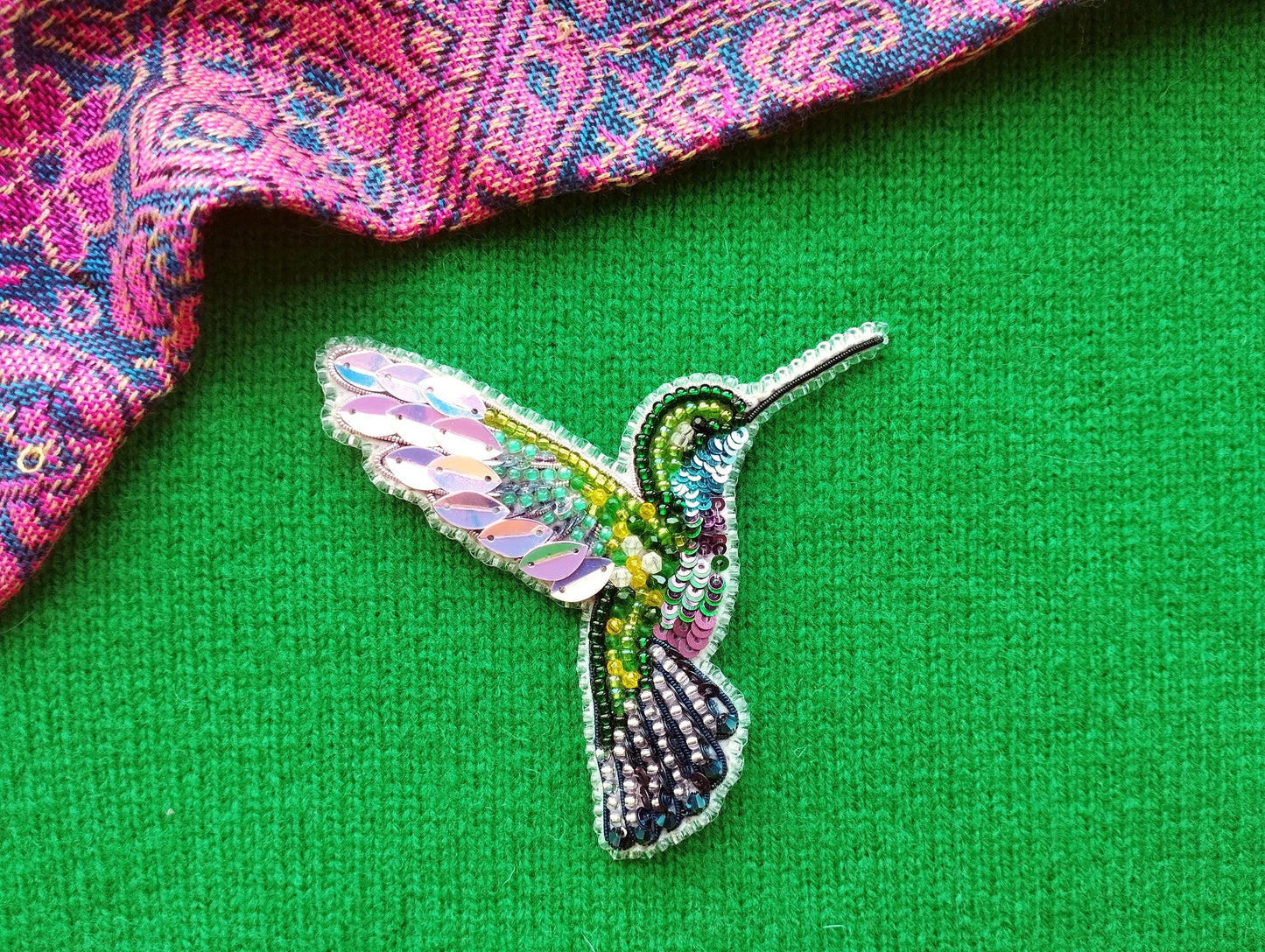 A colorful, intricately designed beaded hummingbird brooch is displayed on a green fabric surface, with a patterned fabric visible in the background.