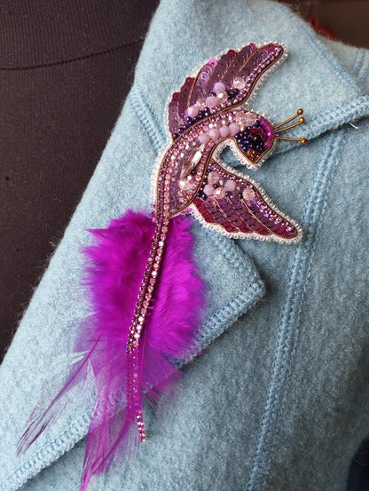 A close-up image of a decorative brooch or pin with a bird-like design, featuring a combination of pink feathers, beads, and a metallic beaded head.