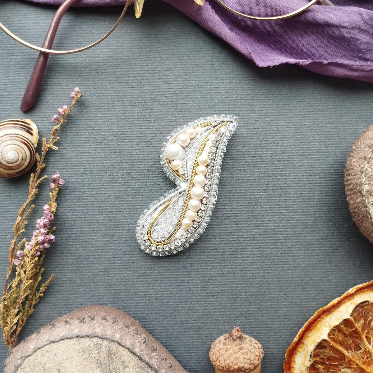 a variety of objects, including a decorative brooch, a pair of glasses, a seashell, a rock, a piece of fruit, and a small acorn, all arranged on a gray surface.