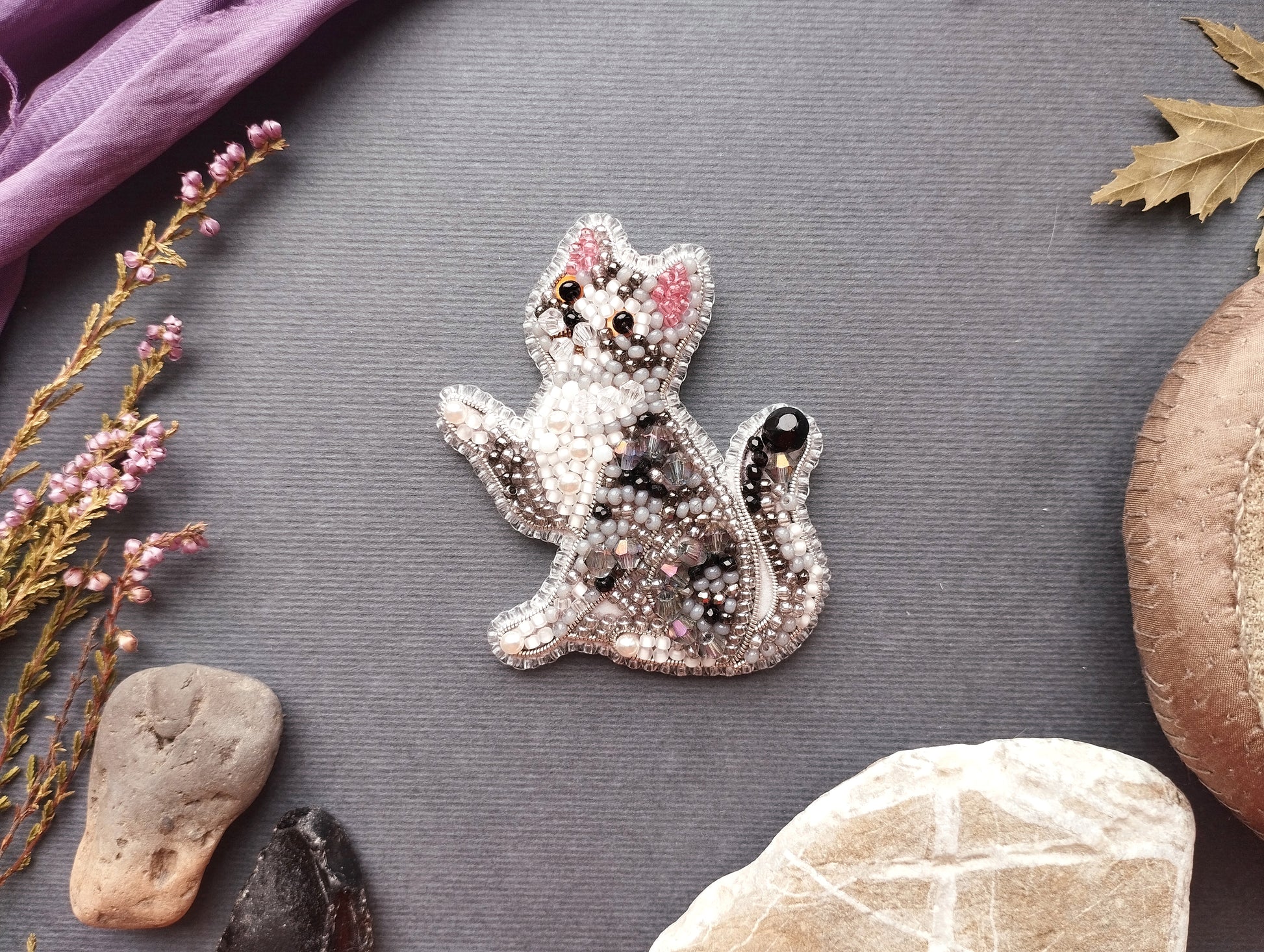 a decorative cat brooch made of beads and surrounded by various natural elements such as leaves, rocks, and flowers.