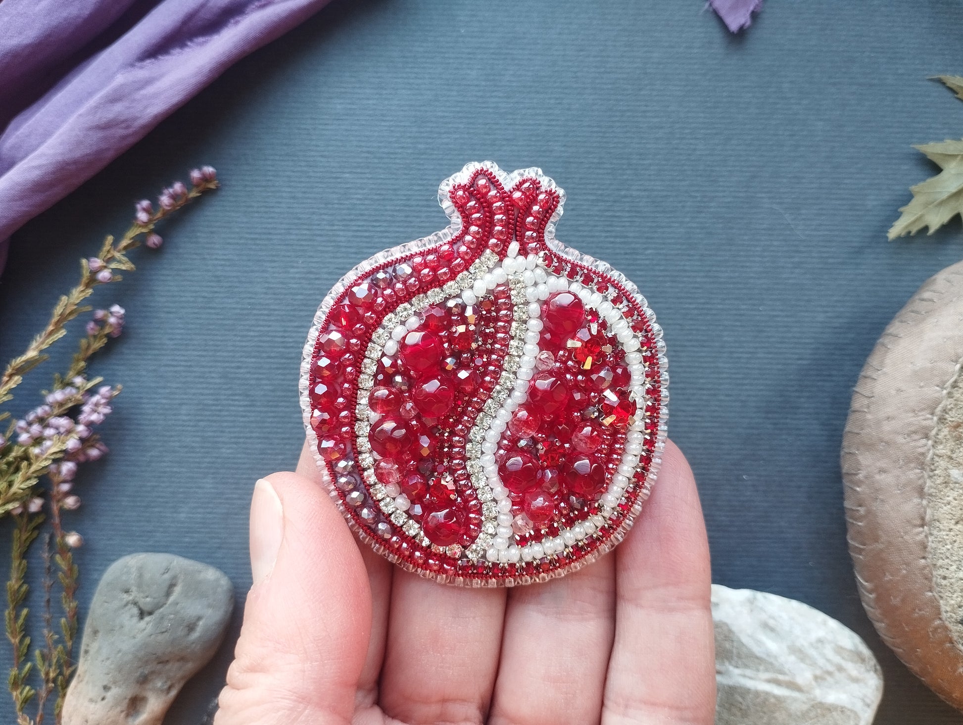 A hand holding a decorative pomegranate-shaped brooch with red and white beads, against a blue background with some purple flowers and rocks.