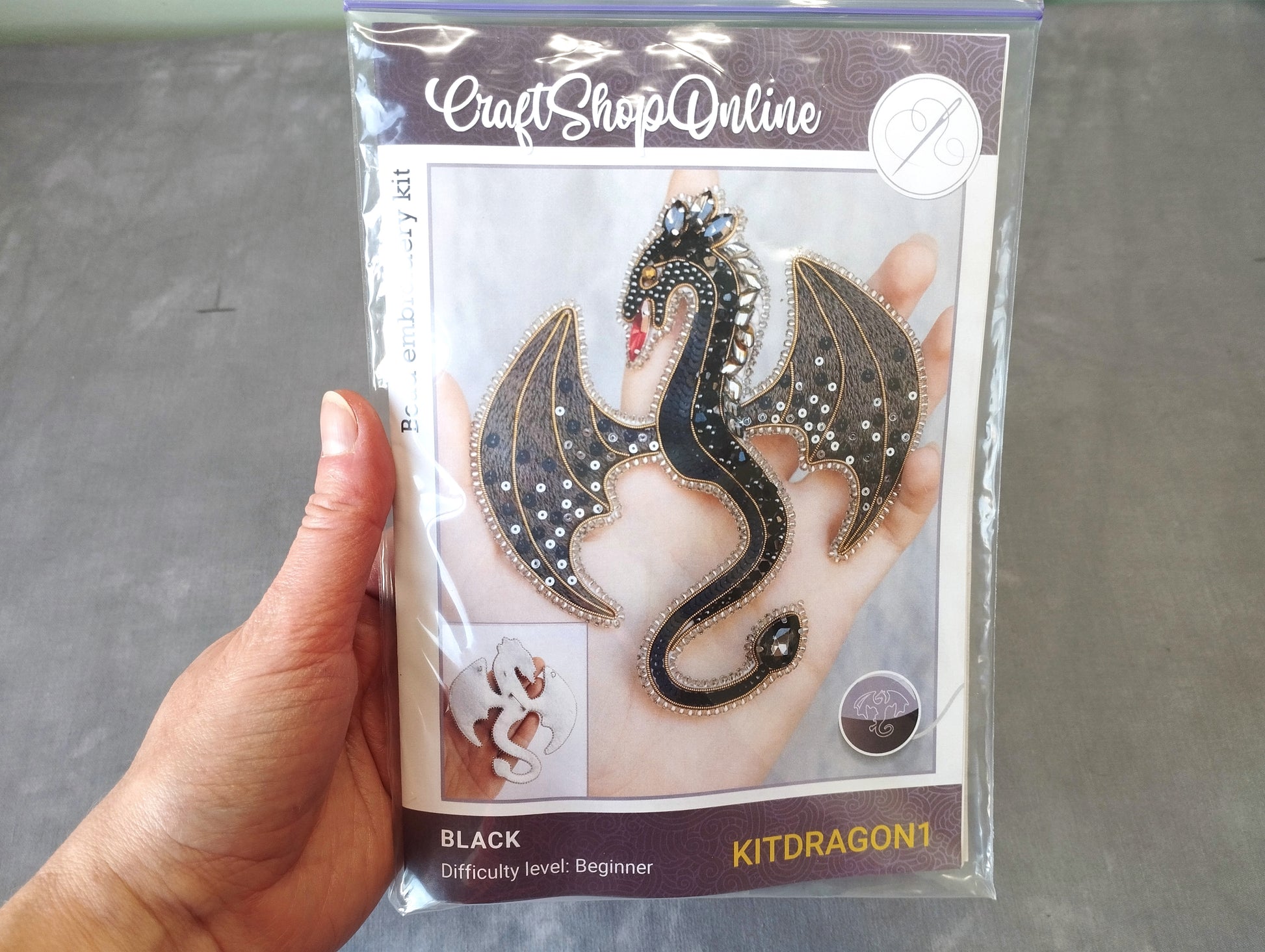 A hand holding a package containing a kit for creating a black dragon-shaped piece of jewelry.