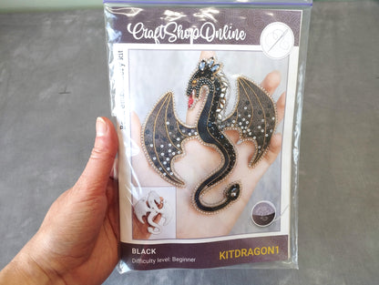 A hand holding a package containing a kit for creating a black dragon-shaped piece of jewelry.
