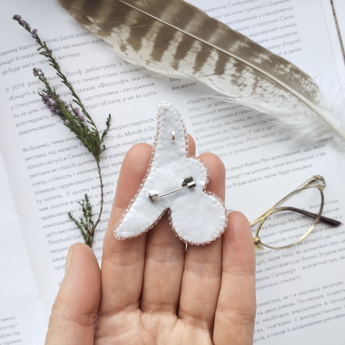 A hand holding a white felt butterfly pin, surrounded by a feather, a plant, and a pair of glasses on a page of text.