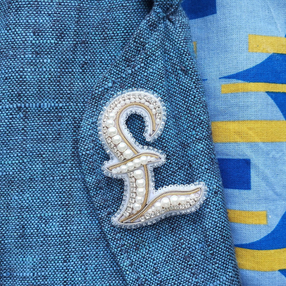 A close-up image of a blue fabric with a decorative monogram made of pearls and rhinestones, placed on a blue fabric with a yellow and blue geometric pattern.