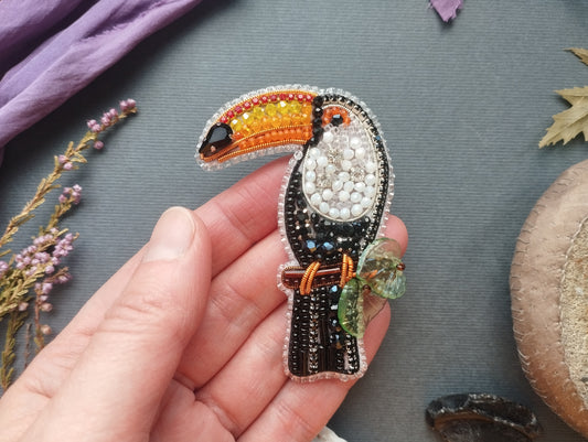 A hand holding a colorful, intricately designed bird brooch against a gray background.