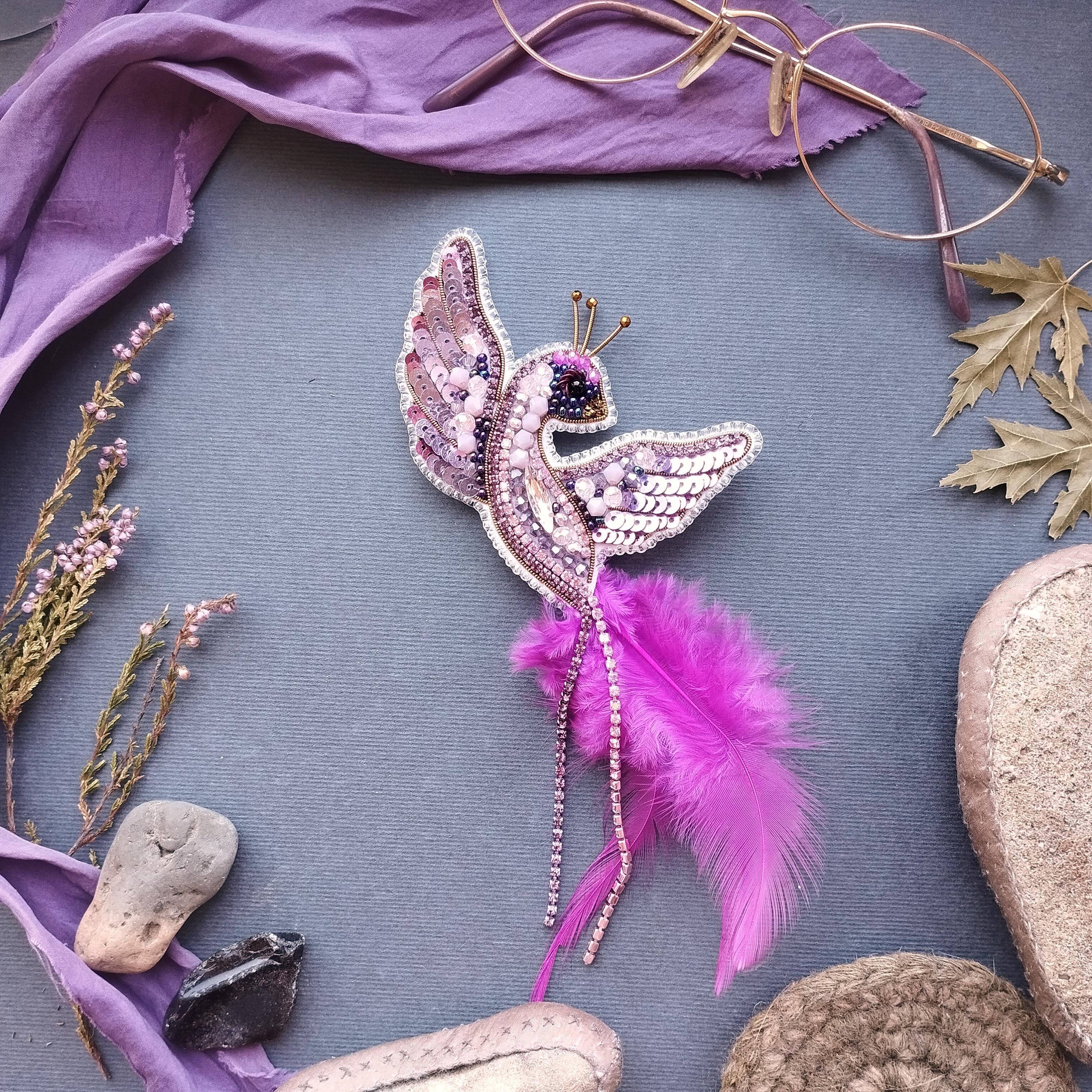 a decorative bird brooch with a pink feather, surrounded by various accessories such as a purple scarf, glasses, and other natural elements like leaves and flowers.