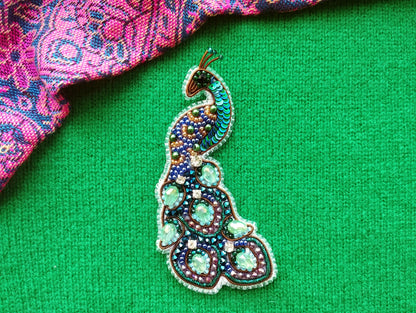 A colorful, intricately designed peacock brooch is displayed against a vibrant green background.