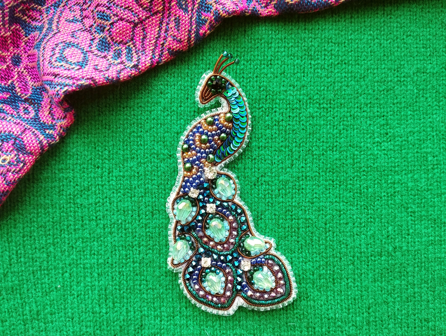A colorful, intricately designed peacock brooch is displayed against a vibrant green background.