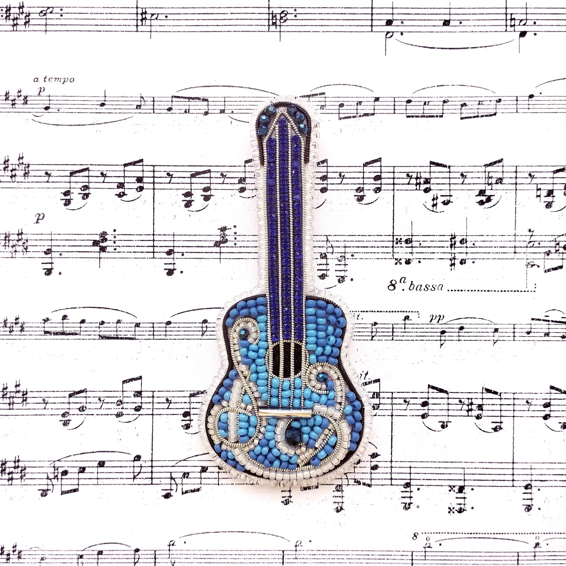 A blue and silver guitar-shaped pin is placed on top of sheet music with musical notes surrounding it.