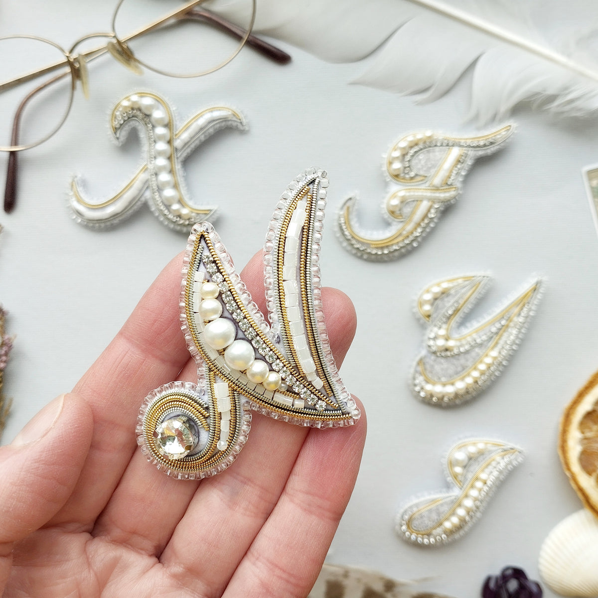 a hand holding a decorative piece of jewelry, which appears to be a beaded or embroidered accessory. The background includes various other decorative elements, such as feathers and pearls, creating a visually appealing and artistic composition.