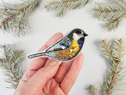 a hand holding a bird brooch with a pine branch in the background
