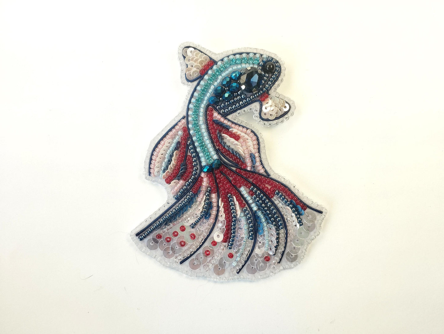 A colorful, intricately designed fish-shaped embroidery piece made of various beads and sequins in shades of blue, red, and pink.