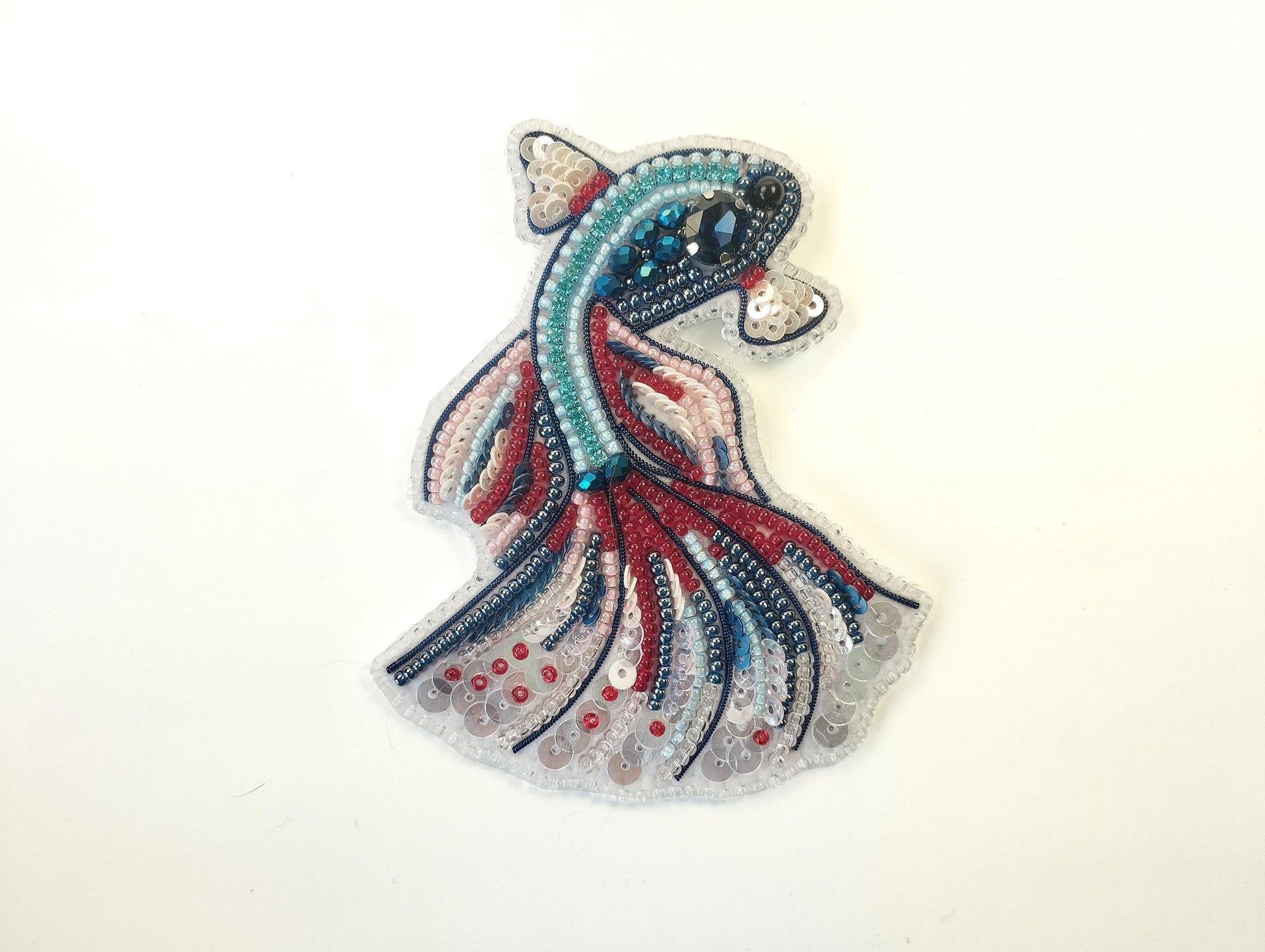 A colorful, intricately designed fish-shaped embroidery piece made of various beads and sequins in shades of blue, red, and pink.