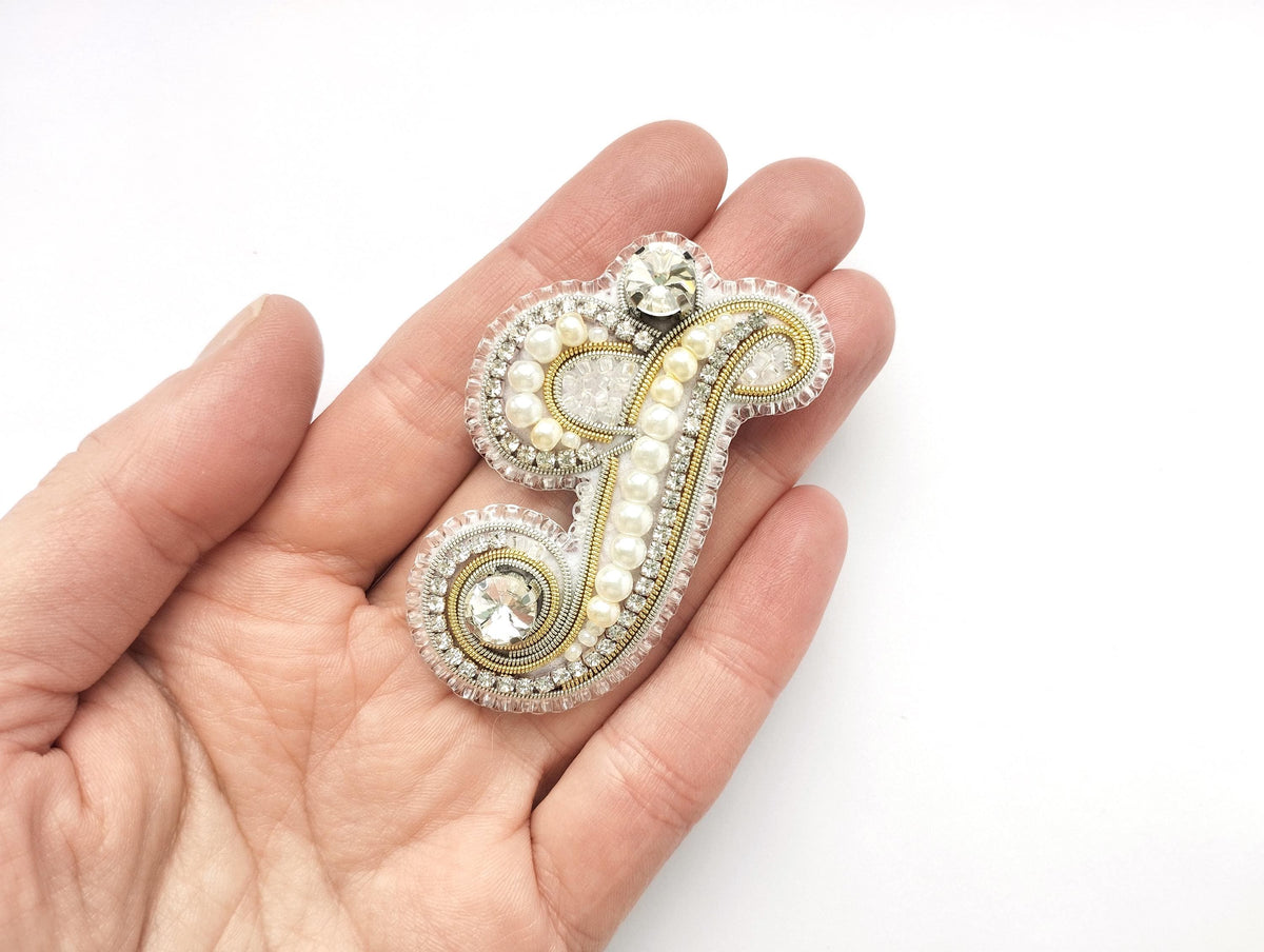A hand holding a decorative piece of jewelry, which appears to be a monogrammed letter or symbol made of various materials such as pearls, crystals, and beads.