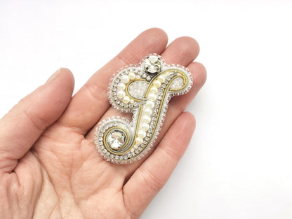 A hand holding a decorative piece of jewelry, which appears to be a monogrammed letter or symbol made of various materials such as pearls, crystals, and beads.