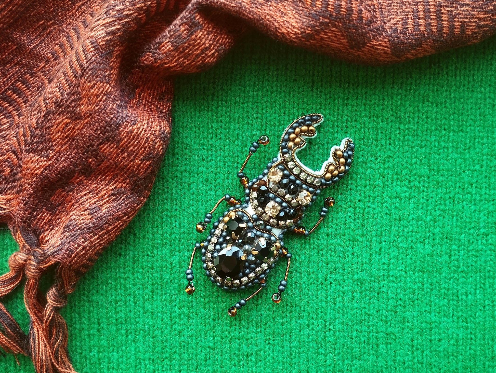 A detailed embroidered or beaded insect brooch is displayed on a green background, with a brown and orange patterned fabric or scarf partially visible to the left.