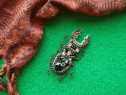 A detailed embroidered or beaded insect brooch is displayed on a green background, with a brown and orange patterned fabric or scarf partially visible to the left.