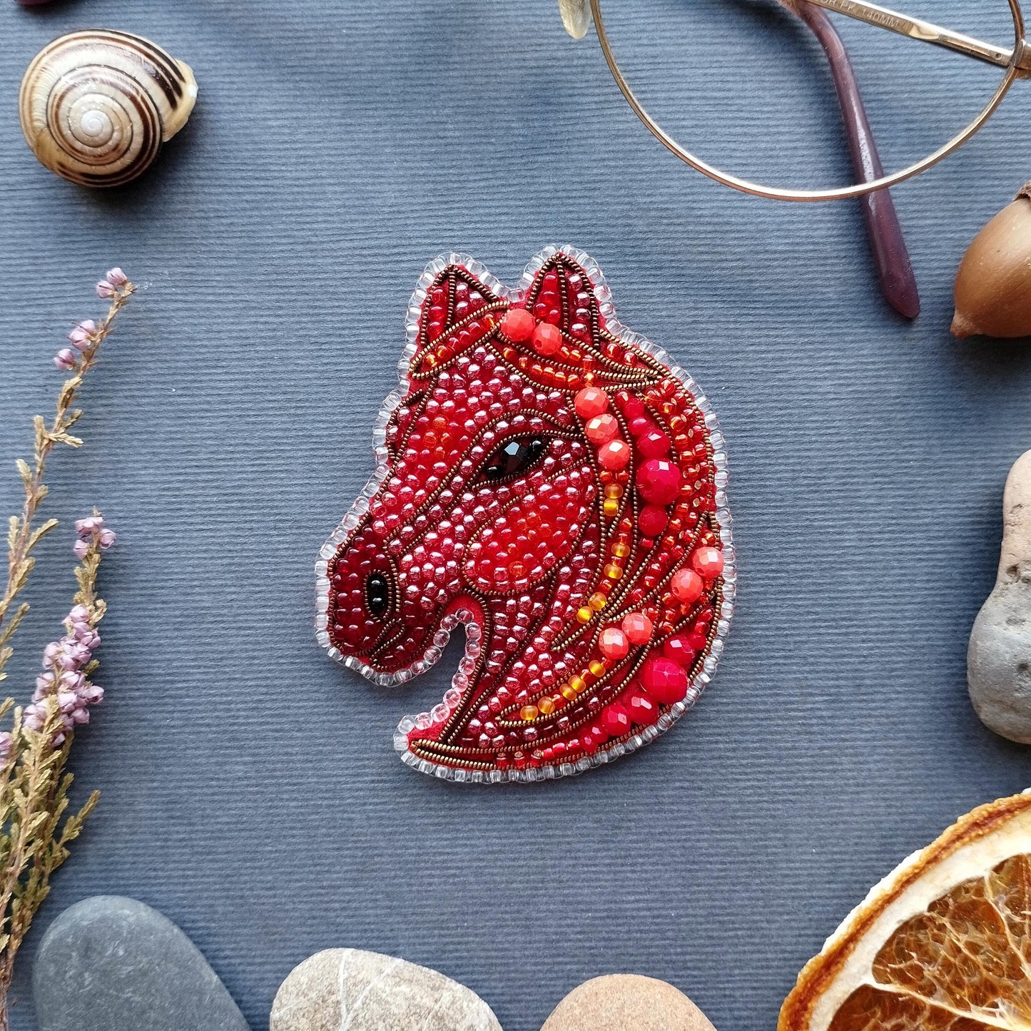 a red beaded embroidered piece of art resembling a horse's head, surrounded by various objects such as a glass, a seashell, a rock, and a slice of orange.