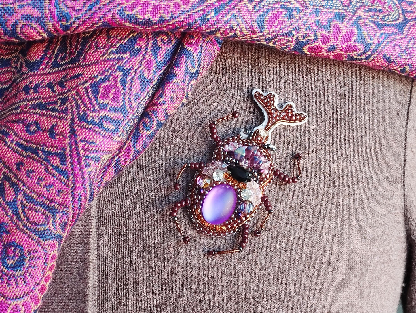 A decorative brooch in the shape of a purple beetle, adorned with jewels and beads, is displayed on a person&#39;s clothing.