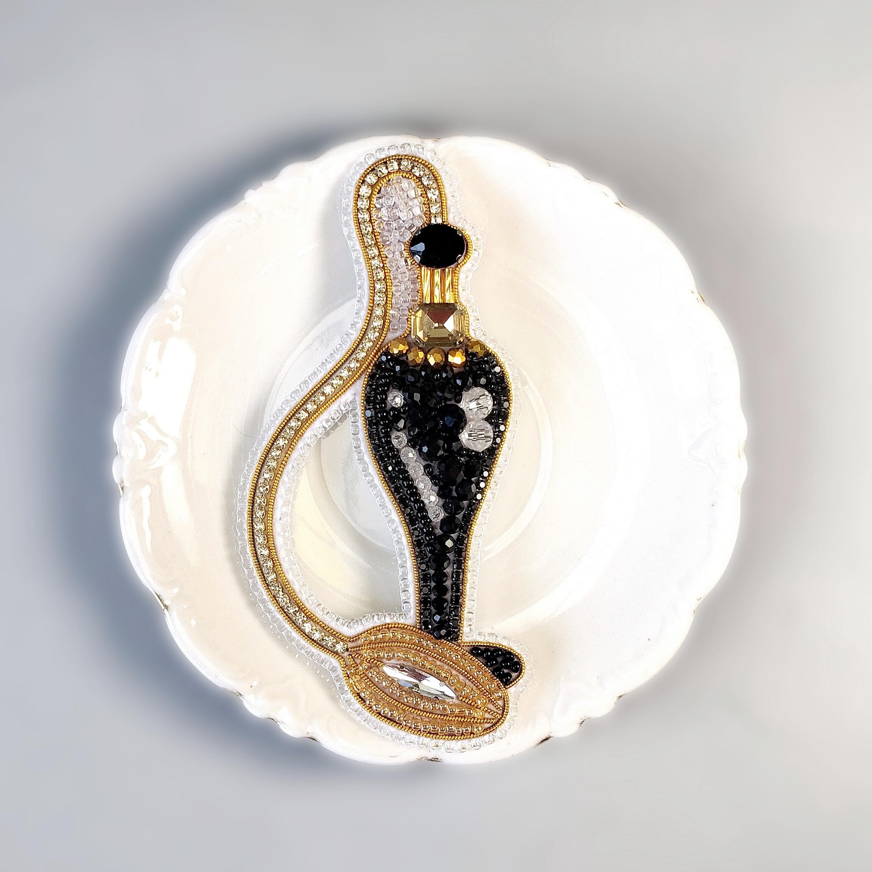 A decorative black and gold perfume bottle with a gold cap and a gold chain is displayed on a white plate against a light background.
