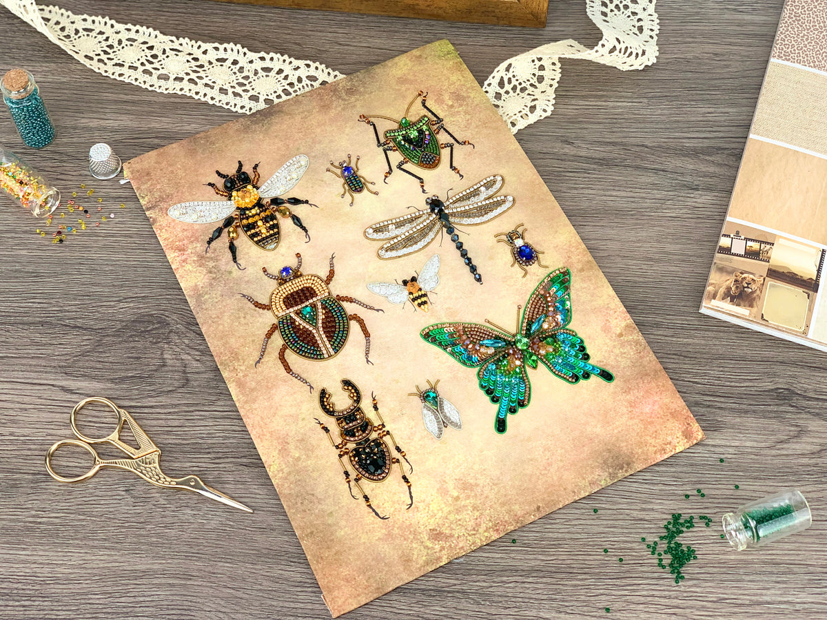 a collection of various insects, including bees, butterflies, and dragonflies, arranged on a piece of paper or card. The insects are intricately detailed and appear to be crafted from beads or embroidery. The scene also includes some crafting supplies, such as scissors and a small bottle, suggesting that the insects are being created or displayed as a work of art or craft project.