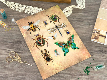 a collection of various insects, including bees, butterflies, and dragonflies, arranged on a piece of paper or card. The insects are intricately detailed and appear to be crafted from beads or embroidery. The scene also includes some crafting supplies, such as scissors and a small bottle, suggesting that the insects are being created or displayed as a work of art or craft project.