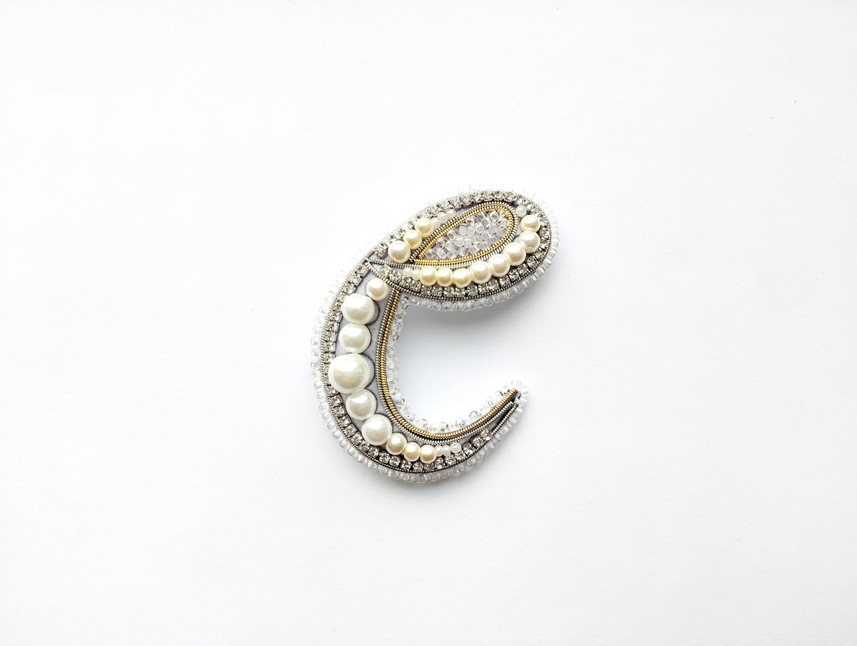 A decorative letter 'C' made of various materials, including pearls, beads, and crystals, against a white background.