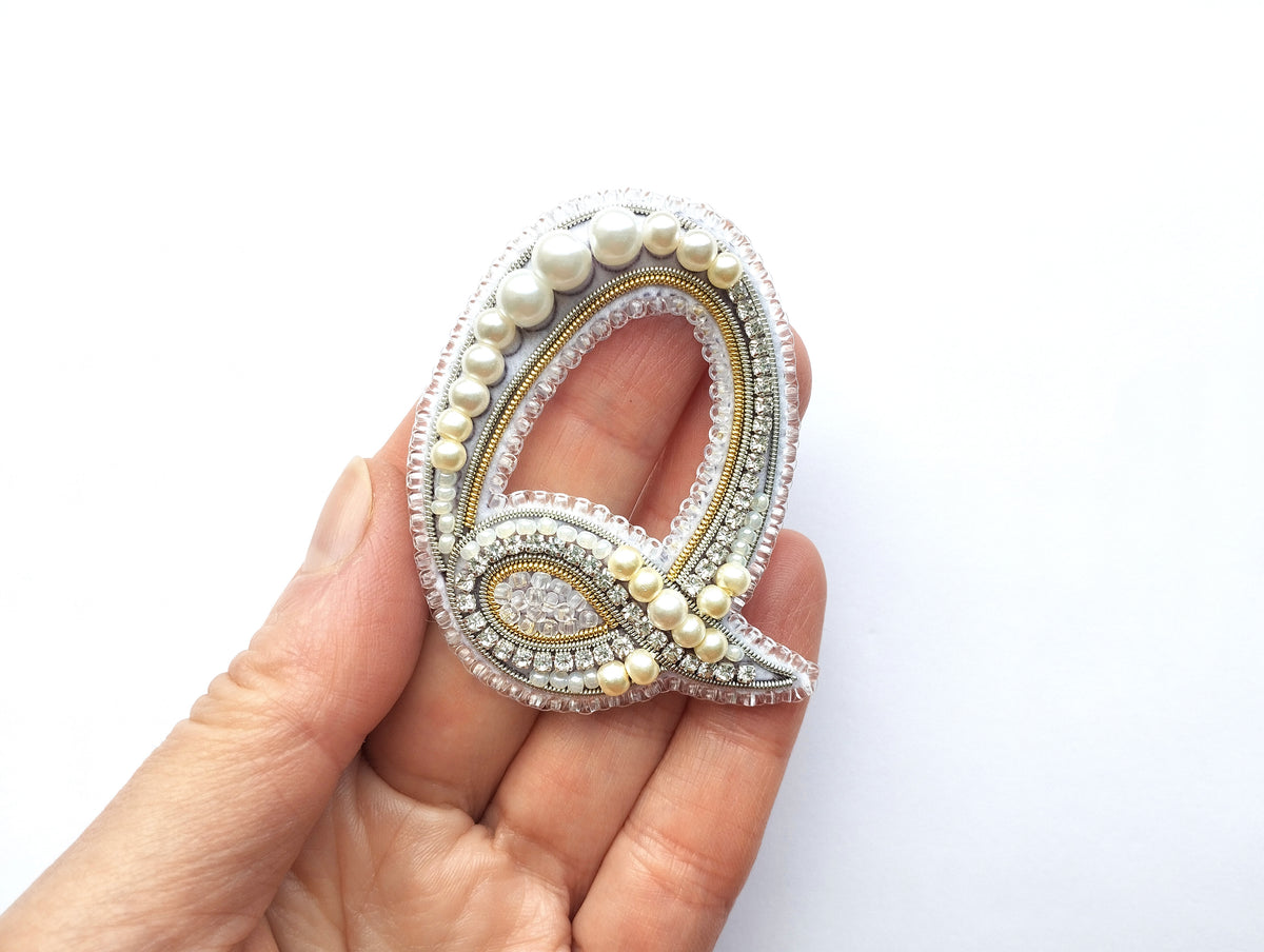 A hand holding a decorative letter 'Q' made of beads, pearls, and other embellishments.